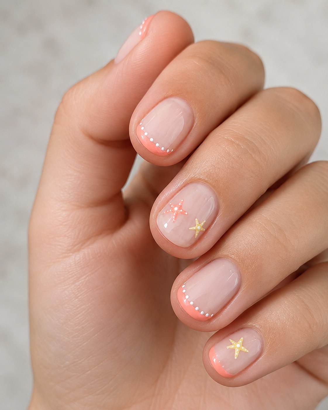 Short shellac nails with a dotted nude french tip, coral edge, and tiny gold starfish charms.